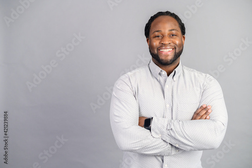 Merry African-American male employee wearing smart casual shirt stands with arms crossed isolated on grey, studio shot of cheerful black businessman, success concept, copy space
