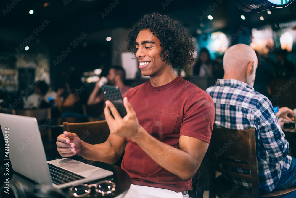 Happy male blogger sharing media files on modern laptop and smartphone technology using bluetooth connection in coworking space, cheerful hipster guy with cellphone browsing netbook information