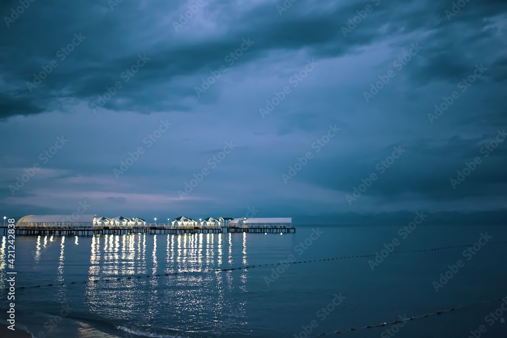 Twilight landscape of pier stretching out into sea. Illumination and umbrellas on ocean bridge. Glare from lamps on water. Nature landscape. Tourism background. Travel and vacation. Cloudy sky.