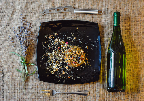 The round head of the French spicy cheese Boulette d'Avesnes sprinkled with different herbs, lies on a black plate. Arrangement: boulette d'avesnes cheese, wine bottle, knife, lavender bouquet photo