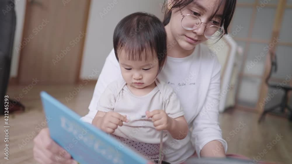 Beautiful asian housewife reading a fairy tales story for her daughter, active smart toddler playing with a doctor stethoscope while sit on mother's lap in living room listening to mom stories Beautiful asian housewife reading a fairy tales story for her daughter, active smart toddler playing with a doctor stethoscope while sit on mother's lap in living room listening to mom stories