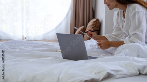 Closeup image of a beautiful young asian woman using laptop computer and working on paperwork while sitting on bed at home