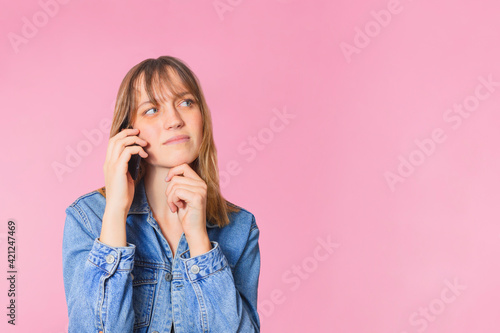 Studio portrait of a woman holding a phone, she is on a call and she is wondering. She has a fringe and blue eyes. She is standing in the center of the image, in front of a pink background. 