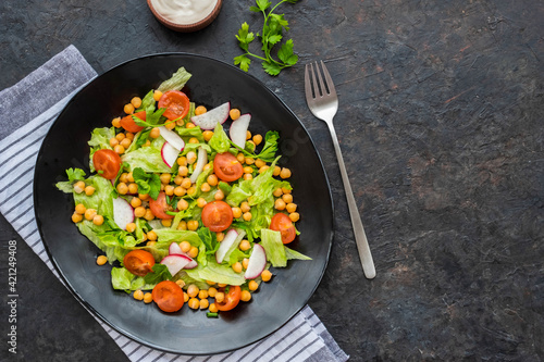 A healthy salad of green lettuce, radishes, tomato with boiled chickpeas on a black plate on a dark concrete background. Served with natural yoghurt sauce. Salad recipes.
