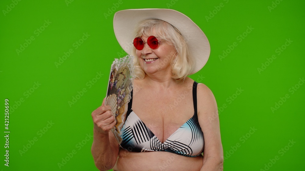 Senior pensioner woman tourist exhales from heat or stuffiness, waves hand fan at herself on chroma key background. Portrait of exited happy old granny traveler in swimsuit model on summer vacations Senior pensioner woman tourist exhales from heat or stuffiness, waves hand fan at herself on chroma key background. Portrait of exited happy old granny traveler in swimsuit model on summer vacations