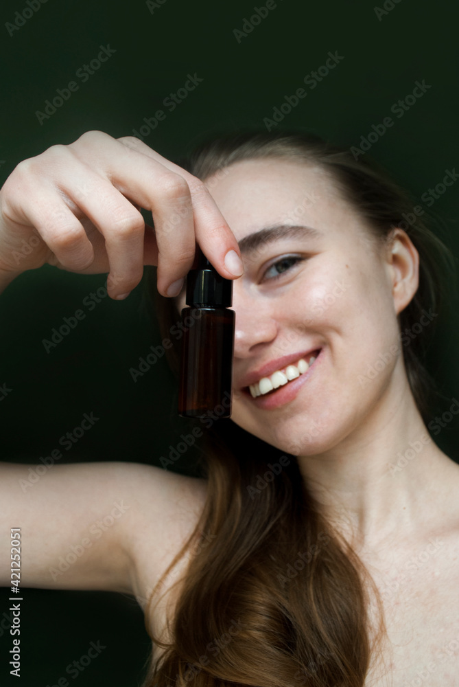 A young smiling blonde shows a bottle of face serum into the camera.Tea tree oil . Selective focus. A young smiling blonde shows a bottle of face serum into the camera.Tea tree oil . Selective focus.