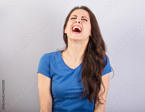 Beautiful positive excited laughting woman with wide opened mouth in blue shirt and long hair on blue background. Closeup photo