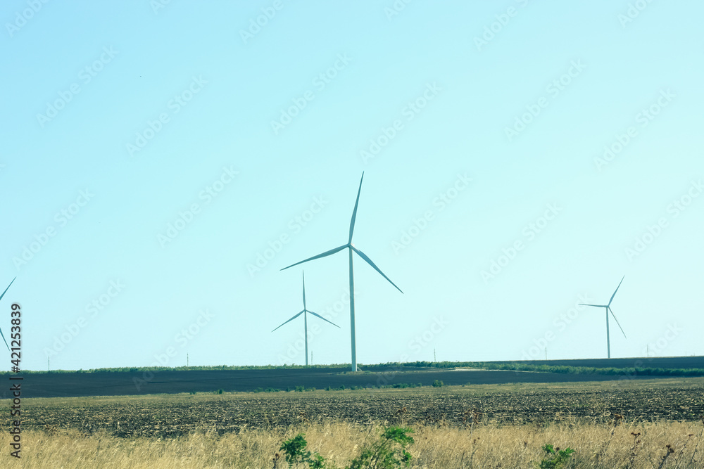 Wind turbines energy converters on wild grass field on sunny day. Local eco friendly wind farm background. Green ecological electricity wallpaper. Modern new environment saving innovation technologies Wind turbines energy converters on wild grass field on sunny day. Local eco friendly wind farm background. Green ecological electricity wallpaper. Modern new environment saving innovation technologies