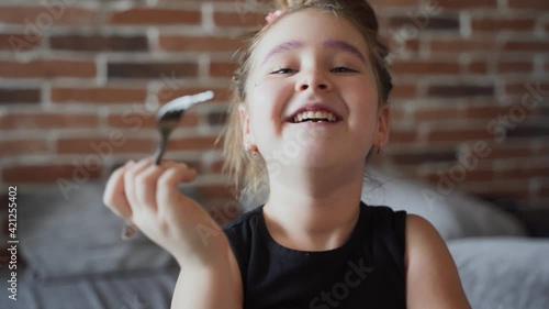 Authentic little girl chews and laughs. She is holding a fork. She smiles and laught. A brick wall is visible in the background. Close up
