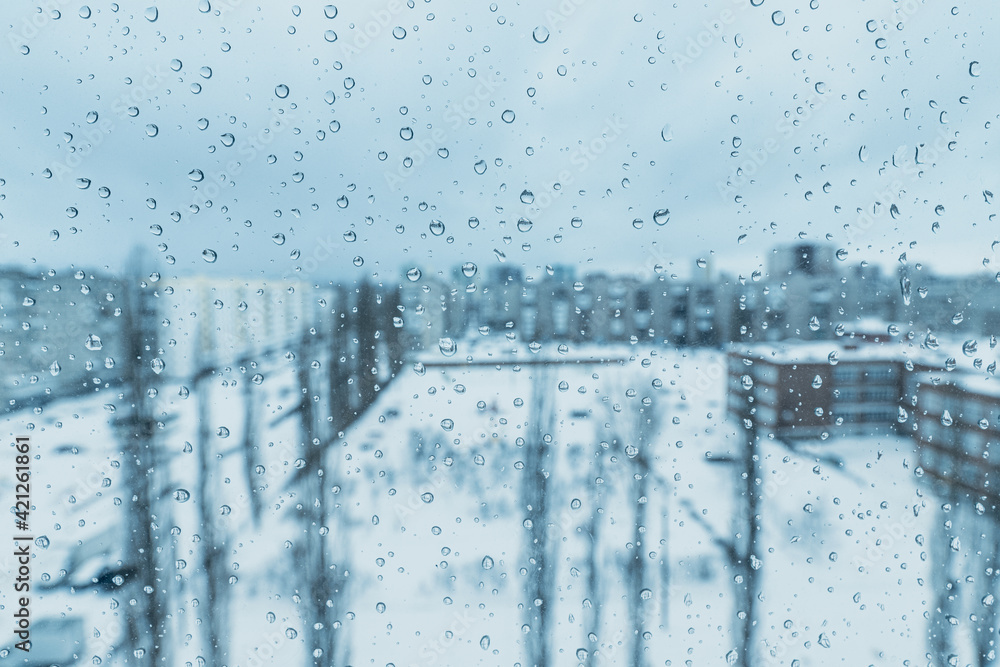 Raindrops on the window with a view of the winter blue snow-covered city. The concept of depression and bad mood