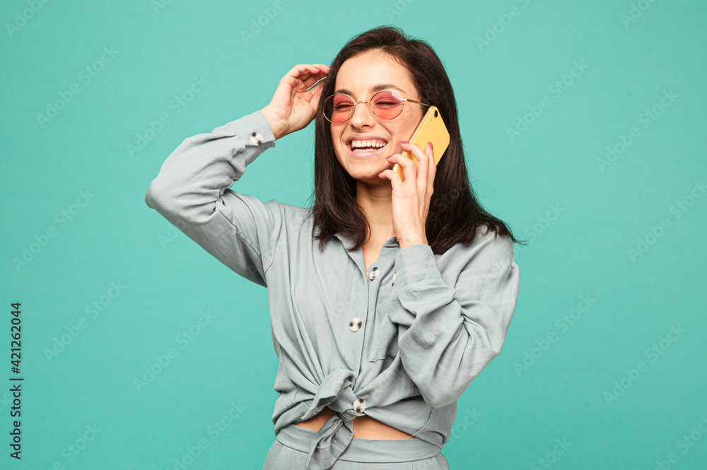Photo of attractive lady in sunglasses talks on phone and smile. Wears grey shirt, isolated turquoise color background Photo of attractive lady in sunglasses talks on phone and smile. Wears grey shirt, isolated turquoise color background