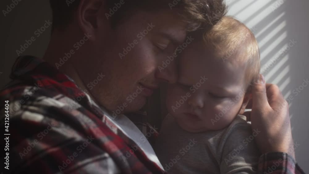 A millennial father holds his son in his arms by the window of the house. The sun's rays from the window shine on the child's face. The family stands and looks out the window. Father hugs son. A millennial father holds his son in his arms by the window of the house. The sun's rays from the window shine on the child's face. The family stands and looks out the window. Father hugs son.
