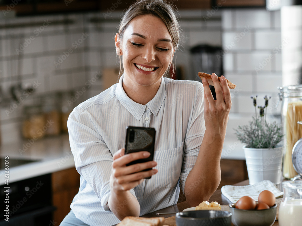 Happy woman eating sandwich in the kitchen. Smiling woman using the phone, reading news online.