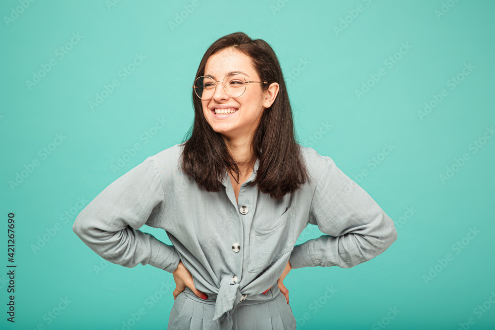 Photo of cute female in glasses smiles nicely. Wears grey shirt, isolated turquoise color background. Photo of cute female in glasses smiles nicely. Wears grey shirt, isolated turquoise color background.