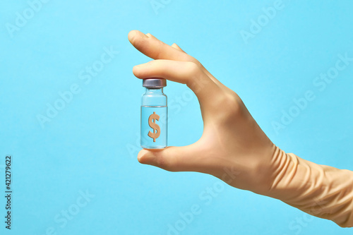 A hand in a medical glove holds a transparent bottle with a vaccine on a blue background. The bottle has a dollar sign. Pharmaceutical companies profit concept.