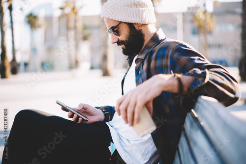 Middle Eastern male blogger in sunglasses and trendy hat checking received email message using 4g internet during leisure in city, hipster guy with coffee to go browsing wireless on smartphone