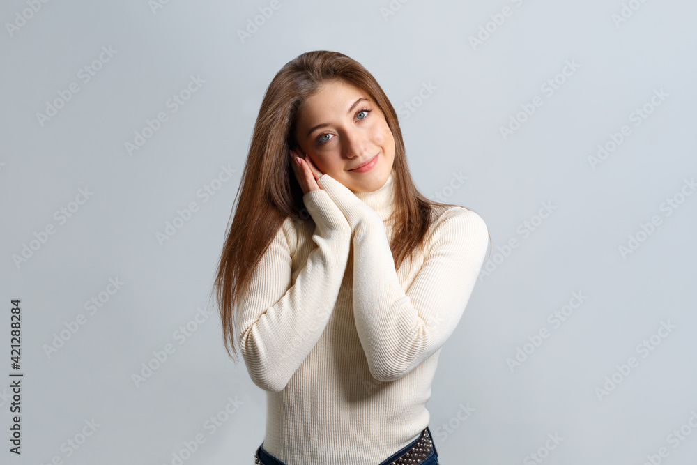 A beautiful girl folded her palms under her cheek for sleep and looks at the camera. Young brown-haired woman stands in a sweater on an isolated gray background. A beautiful girl folded her palms under her cheek for sleep and looks at the camera. Young brown-haired woman stands in a sweater on an isolated gray background.