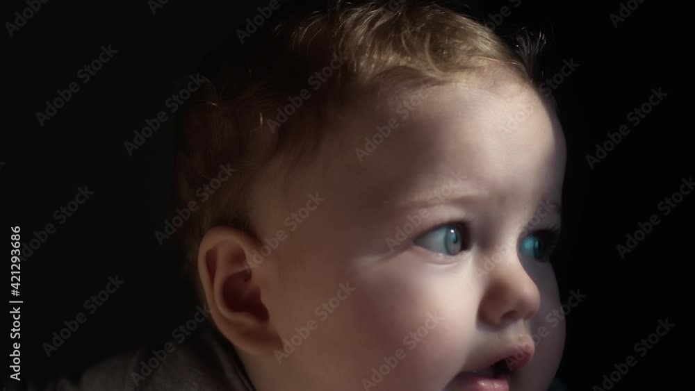 The baby is in the arms of his mother, looks with blue eyes. Millennial mother holds her son in her arms by the window of the house. The child's face in the sun. Close-up portrait The baby is in the arms of his mother, looks with blue eyes. Millennial mother holds her son in her arms by the window of the house. The child's face in the sun. Close-up portrait