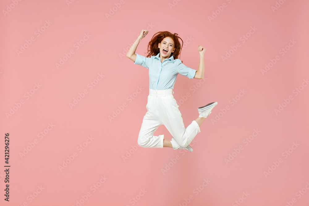 Full length young fun student successful overjoyed happy redhead woman 20s in blue shirt pants do winner gesture clench fist celebrate jumping high isolated on pastel pink background studio portrait Full length young fun student successful overjoyed happy redhead woman 20s in blue shirt pants do winner gesture clench fist celebrate jumping high isolated on pastel pink background studio portrait