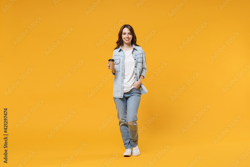 Full length of young smiling friendly fun student happy woman 20s in casual denim shirt white t-shirt holding paper cup of coffee drinking hot tea isolated on yellow color background studio portrait Full length of young smiling friendly fun student happy woman 20s in casual denim shirt white t-shirt holding paper cup of coffee drinking hot tea isolated on yellow color background studio portrait