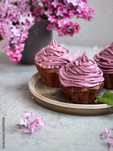 Baking, lilac, spring. Delicious chocolate cupcakes with lilac cream on top. Filled with chocolate. Surrounded by fresh lilacs. On a concrete countertop. Gray background. Space for the text. Content.