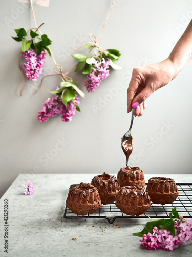 Baking, lilac, spring. Chocolate cupcakes with lilac cream on top. Women's hands pour chocolate paste over pastries. Surrounded by fresh lilacs. On a concrete countertop. Gray background. Copy paste.