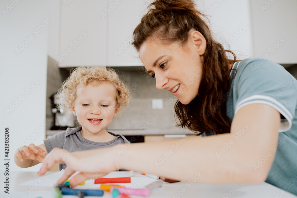 A beautiful mother sits at the kitchen table with her preschool-aged daughter and makes something out of plasticine. Mom is spending her afternoon free time with her daughter. A beautiful mother sits at the kitchen table with her preschool-aged daughter and makes something out of plasticine. Mom is spending her afternoon free time with her daughter.
