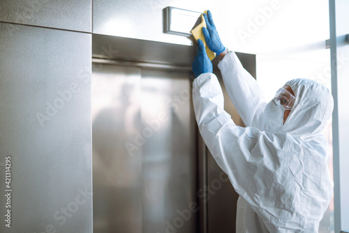 Disinfecting  of the elevator. Man in a protective suit and mask sprays disinfectants buttons of the lift. Protection agsinst COVID-19 disease. Cleaning concept.  © maxbelchenko