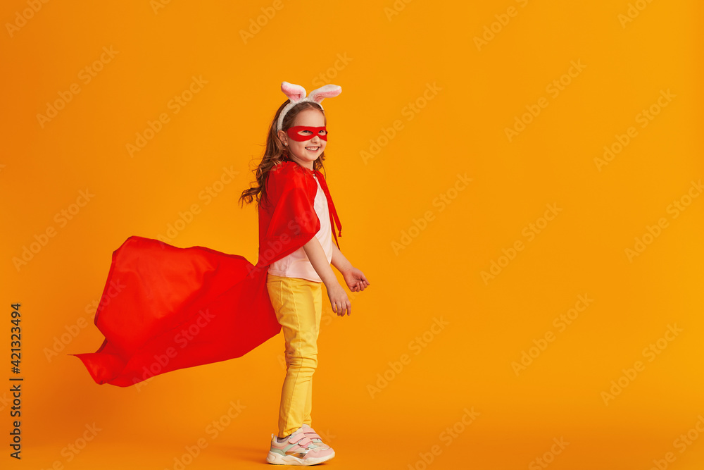 fun little girl is playing in superhero costume. baby is dressed in rabbit ears, red cape, mask, holding out her hand on yellow background. concept girl's superpowers, feminism, striving for victory