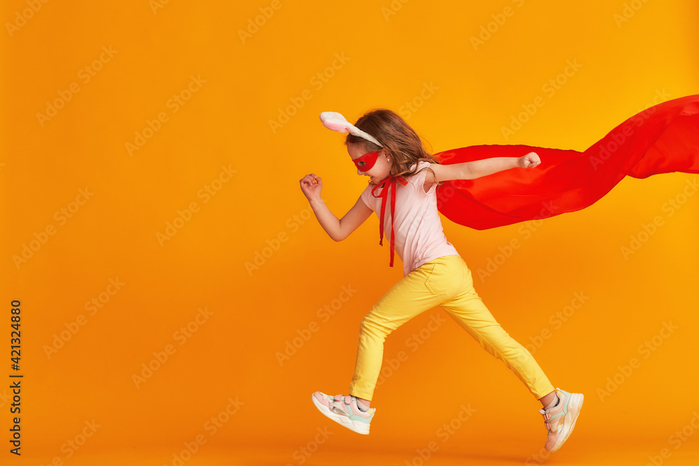 fun little girl is playing superhero game. baby is dressed in rabbit ears, red cape and mask, reaches out and runs on yellow background. concept girl's superpowers, feminism, striving for victory fun little girl is playing superhero game. baby is dressed in rabbit ears, red cape and mask, reaches out and runs on yellow background. concept girl's superpowers, feminism, striving for victory