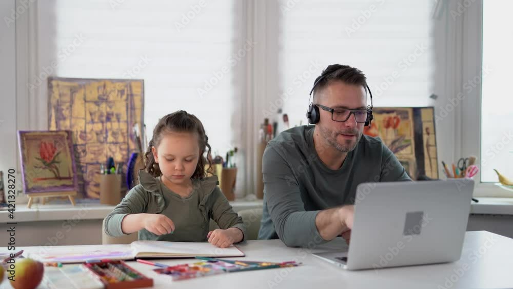 Young caucasian family busy working in their room at the table. Child girl drawing next to dad who is working online with laptop and headphones. Babysitting during covid-19 lockdown. Front view. Young caucasian family busy working in their room at the table. Child girl drawing next to dad who is working online with laptop and headphones. Babysitting during covid-19 lockdown. Front view.