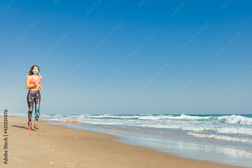 Girl running sport in the beach with covid mask and a blue sky and a blue sea with whit waves.