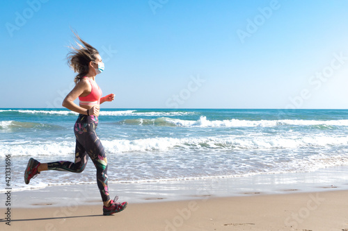 Girl running sport in the beach with covid mask and a blue sky and a blue sea with whit waves.