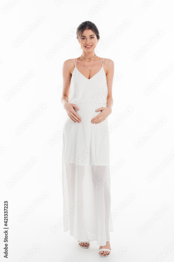 full length view of happy, pregnant fiancee touching belly while smiling at camera on white full length view of happy, pregnant fiancee touching belly while smiling at camera on white