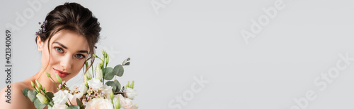 happy fiancee looking at camera while holding wedding bouquet isolated on grey, banner