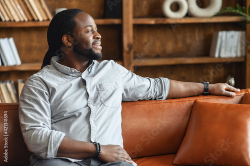 Portrait of a young hispanic black man resting on sofa, thinking about the future, handsome positive man with hand on the back of the sofa sitting in living room, daydreaming and relaxing at home