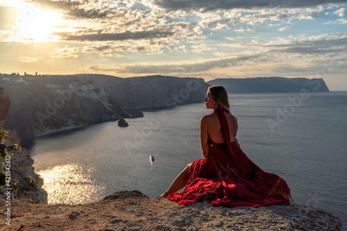 A girl with loose hair in a red dress sits on a rock rock above the sea. In the background, the sun rises from behind the mountains.