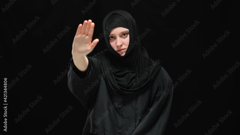 Scared stressed Muslim woman in hijab raising hand to camera. Portrait of terrified young beautiful victim of tyranny posing at black background. Scared stressed Muslim woman in hijab raising hand to camera. Portrait of terrified young beautiful victim of tyranny posing at black background.