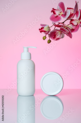 Hand cream in a bottle and jar on a pink background with a reflection. Pink orchid in a top right corner.