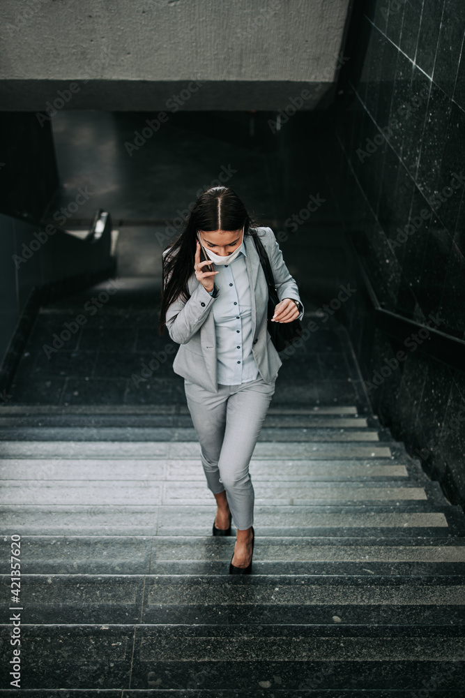 Elegant and worried business woman using smartphone and standing on empty city street. She is wearing protective mask to protect herself from dangerous flu or virus. Corona virus or Covid-19 concept.