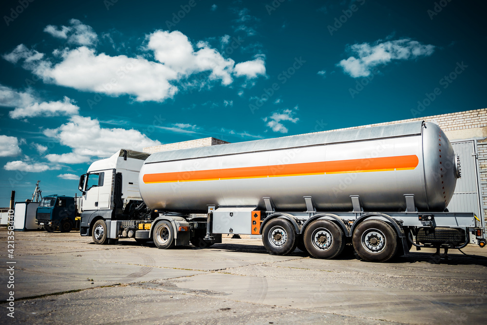 Truck with trailer, tank with flammable liquid, sunny day outside, metallic color container, blue sky with white clouds, gravel