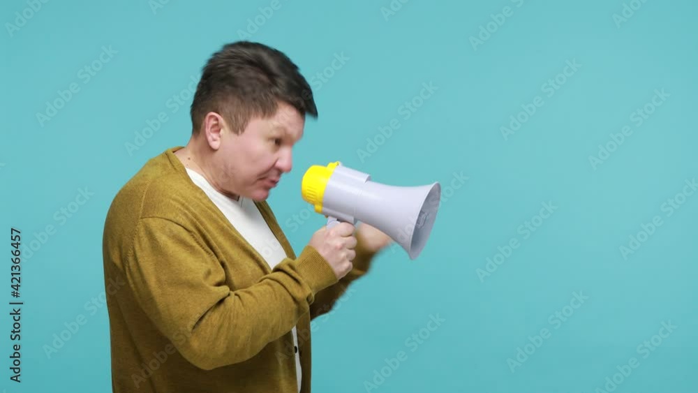 Profile portrait concentrated mature man leader in white t-shirt and cardigan talking at megaphone, expressing his point of view, making announcement. Indoor studio shot isolated on blue background Profile portrait concentrated mature man leader in white t-shirt and cardigan talking at megaphone, expressing his point of view, making announcement. Indoor studio shot isolated on blue background