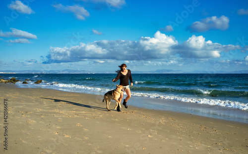 Happy woman and dog running on beach at sunset. Young woman exercising with her German Shepherd Dog as they run side by side along a sandy tropical beach with gentle surf and calm ocean.