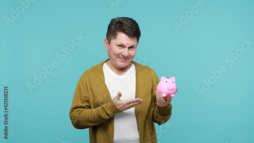 Smiling positive mature dark haired man in white t-shirt and cardigan holding piggy bank showing thumbs up gesture, satisfied with bank deposit, credit. Indoor studio shot isolated on blue background photo