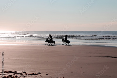 Two girls on their bikes with surfboard by the sand on the beach.