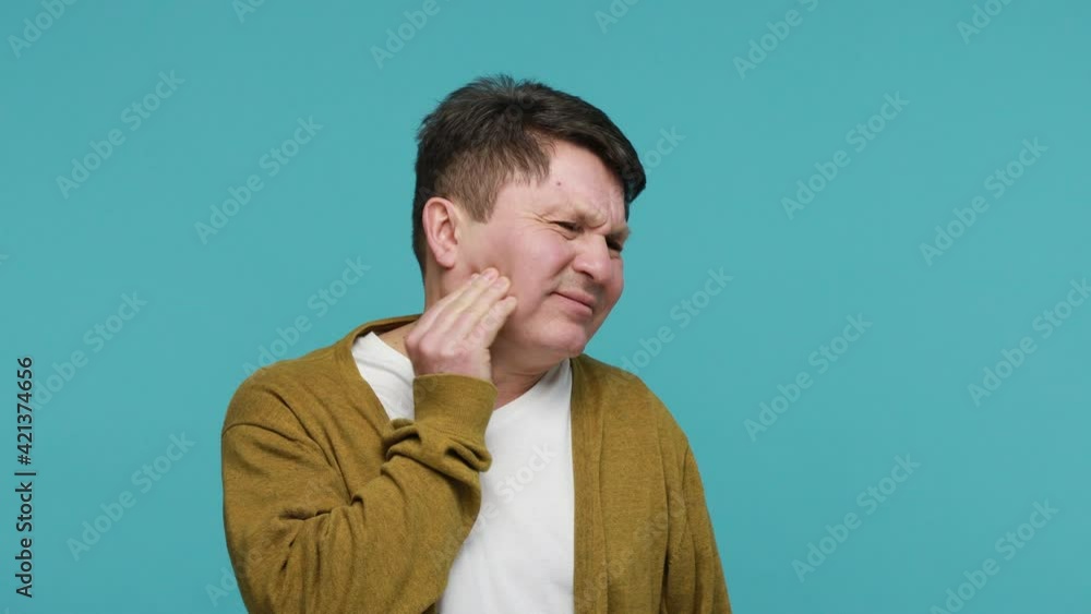 Teeth pain. Sick adult dark haired man in white t-shirt and cardigan rubbing cheek feeling horrible suffer from toothache, dental problems, gingivitis. Indoor studio shot isolated on blue background Teeth pain. Sick adult dark haired man in white t-shirt and cardigan rubbing cheek feeling horrible suffer from toothache, dental problems, gingivitis. Indoor studio shot isolated on blue background