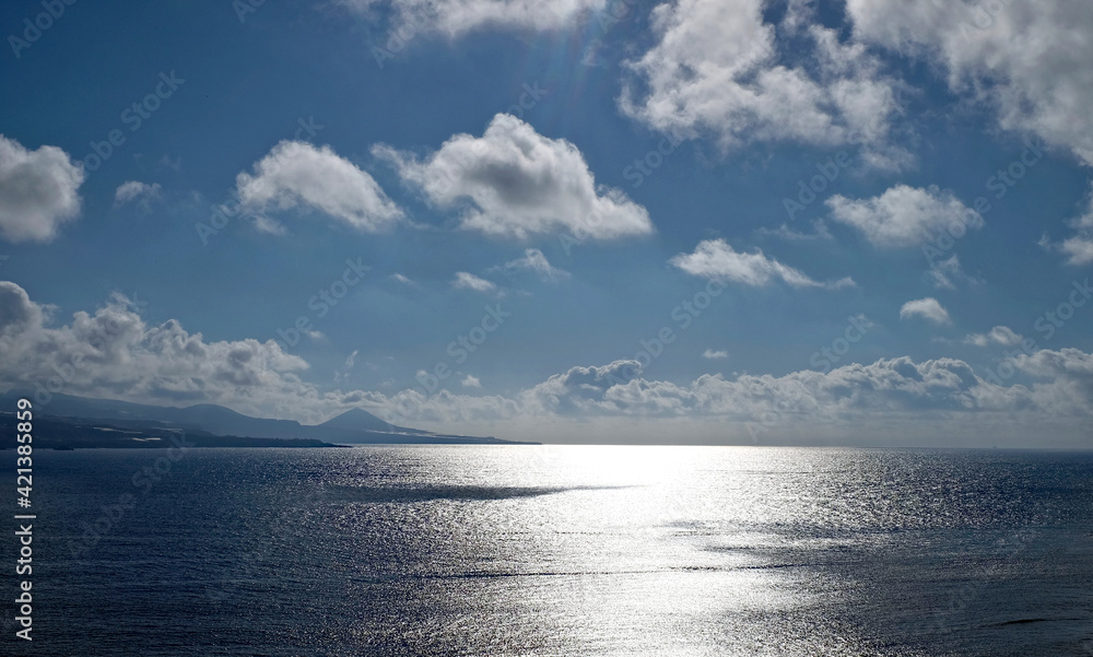 Sea landscape in contrasty bluish light, sun reflected in large open water and remote volcanic slopes of an Atlantic island.