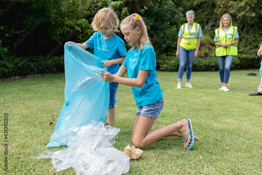 Naklejka premium Caucasian boy and girl with volunteer group kneeling collecting plastic rubbish in field