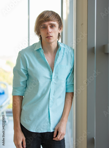 Portrait vertical photo of caucasian adult young man wearing blue shirt look at camera standing in room do look worry, sick and sad and uncomfortable look like patient with bed in background on day