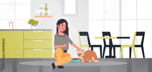 woman feeding hungry cat girl giving her kitty dry food granules domestic life with pet concept modern kitchen interior horizontal full length
